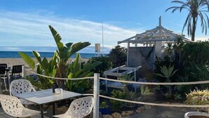 A wooden deck with a table and chairs, overlooking a beach with palm trees and a gazebo. - Bright and airy apartment in Puerto Banus (Marbella)