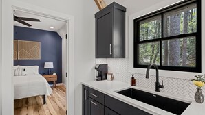 Eine moderne Küche mit einem schwarzen Spülbecken, einer weißen Arbeitsplatte und einem Fenster mit Blick auf einen Wald.