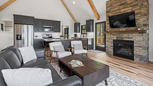 Ein modernes Wohnzimmer mit Kamin aus Naturstein, einem Flachbildfernseher und einem Essbereich, im Hintergrund ist eine Küche zu sehen.