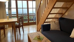 A cozy room with a wooden staircase, a sofa, a dining table, and a view through large glass doors. - Vacation apartment Kampenwand (Rimsting)