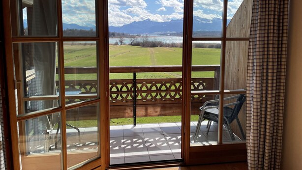 A room with a large sliding glass door leading to a balcony with a view of a mountain and a lake. - Vacation apartment Kampenwand (Rimsting)