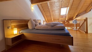 A loft bedroom with a wooden ceiling, a bed with pillows, a bedside table with a lamp, and a fan. - Vacation apartment Kampenwand (Rimsting)