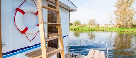 Un bateau équipé d’une échelle en bois, d’une bouée de sauvetage, et offrant une vue sur une rivière bordée d’arbres et où l’on peut apercevoir un troupeau de bovins.