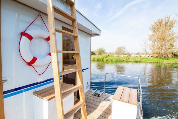 A boat with a wooden ladder, a lifebuoy, and a view of a river with trees and a herd of cattle. - Fully-Equipped Boat 'Lisa' with Terraces (Priepert)