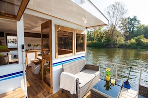 A boat interior with a dining area, a seating area with a table and chairs, and a view of the river. - Fully-Equipped Boat 'Lisa' with Terraces (Priepert)
