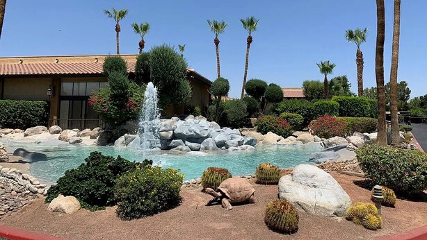 Un jardín con arreglos paisajísticos, una fuente, palmeras y un edificio con techo de tejas.