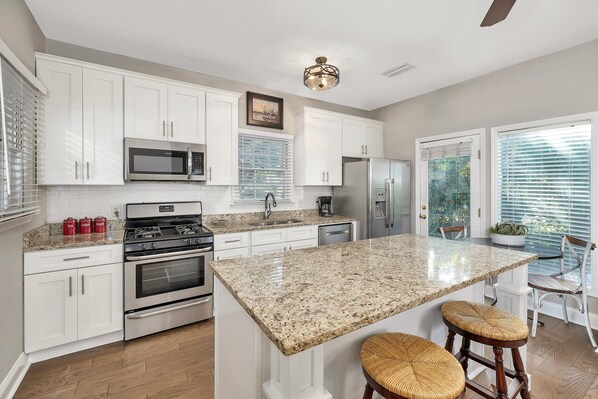 A modern kitchen with white cabinetry, stainless steel appliances, a granite countertop, and a central island with stools. - 1112 Ocean Boulevard (Saint Simons Island)