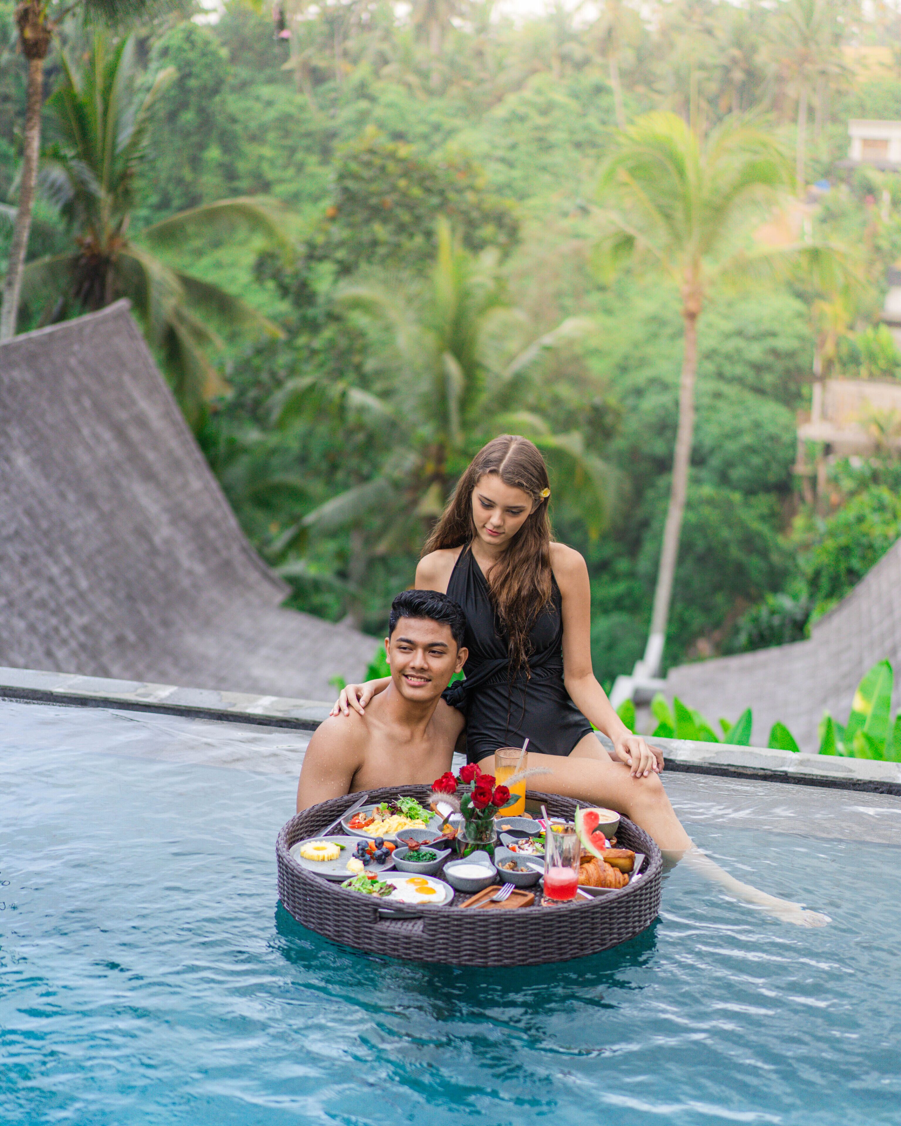 A couple sitting on a floating dining table in a pool surrounded by lush greenery.