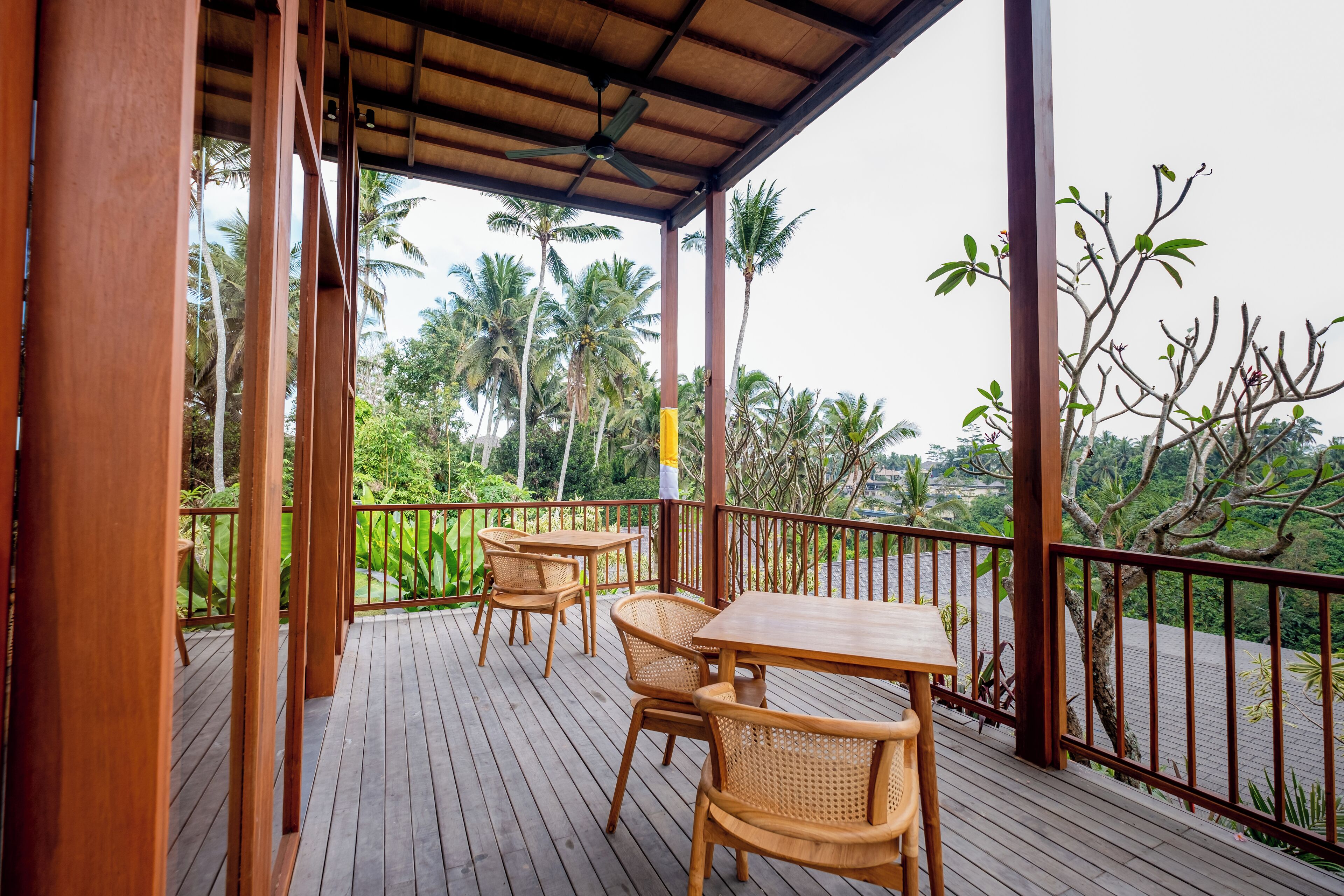 Une terrasse en bois, agrémentée de tables et de chaises, entourée d’une rambarde et offrant une vue sur des palmiers et une végétation luxuriante.