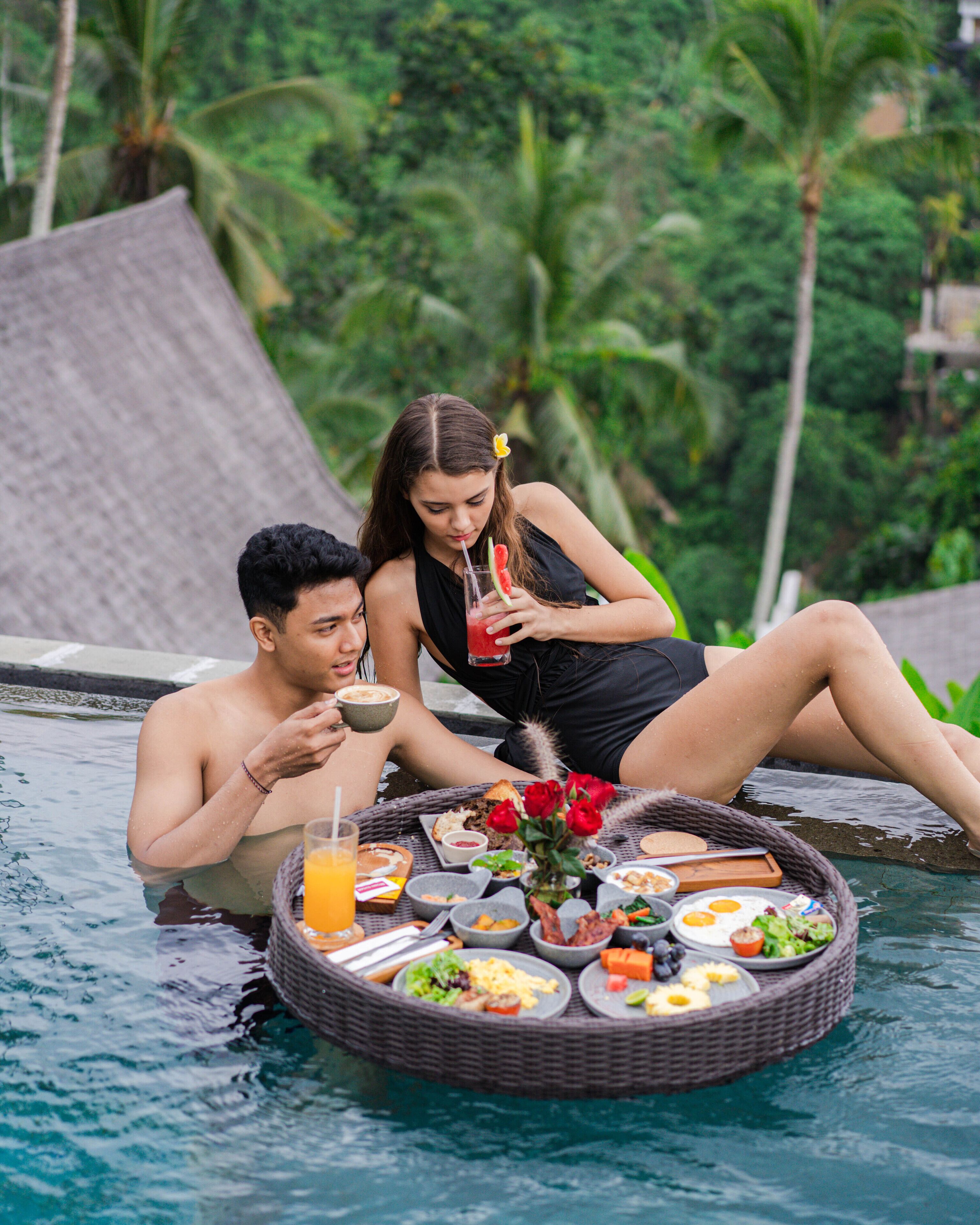 A couple relaxing by a poolside table with a meal and drinks.