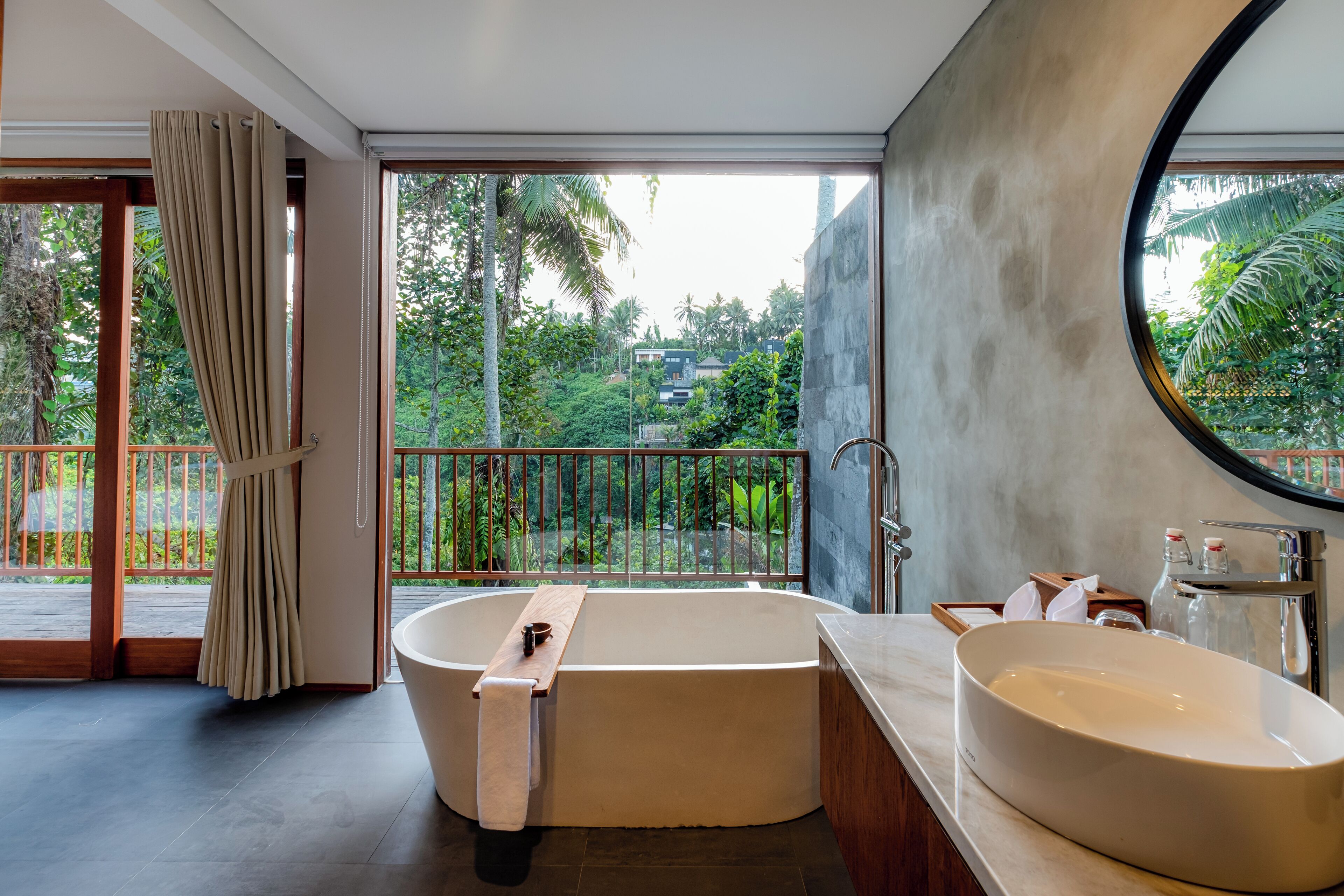 Un baño moderno con una bañera independiente, un lavabo ovalado grande y vistas a exuberante follaje a través de amplios ventanales.