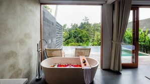 A modern bathroom with a freestanding bathtub, a glass shower enclosure, and a view of a lush garden and pool. - LeRosa Valley Resort (Ubud)
