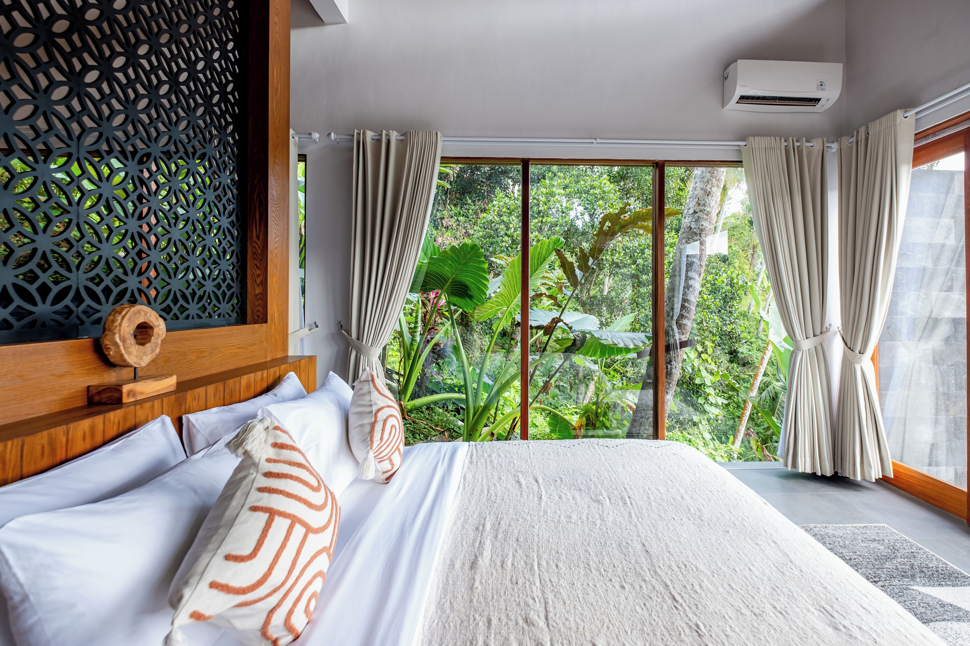 A bedroom with a large bed, a wooden headboard, a decorative wall panel, and a view of lush greenery through large windows.