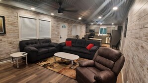 Un salon moderne avec un canapé d’angle, un fauteuil en cuir et une petite table basse. On aperçoit, au fond, une cuisine équipée d’appareils électroménagers en acier inoxydable.