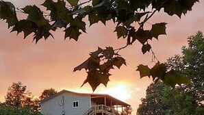 Un chemin de gravier mène à une maison dotée d’un porche, entourée de verdure, avec un coucher de soleil en arrière-plan.