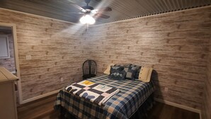 Une chambre avec un mur en bois, un lit recouvert d’une couverture à carreaux, un ventilateur de plafond et une fenêtre avec des stores.