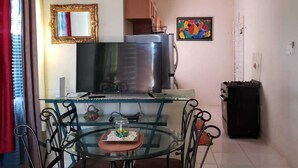 A dining area with a glass table and metal chairs, a television, and a kitchen area with a stove and cabinets. - Jacaranda Vacation Home (Spanish Town)