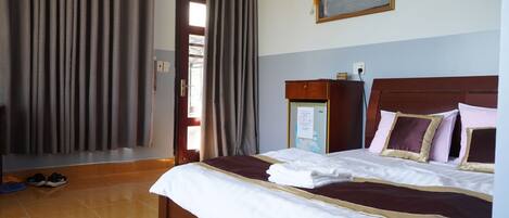 A hotel room with a large bed, a wooden headboard, a nightstand, a painting on the wall, and a door with curtains.