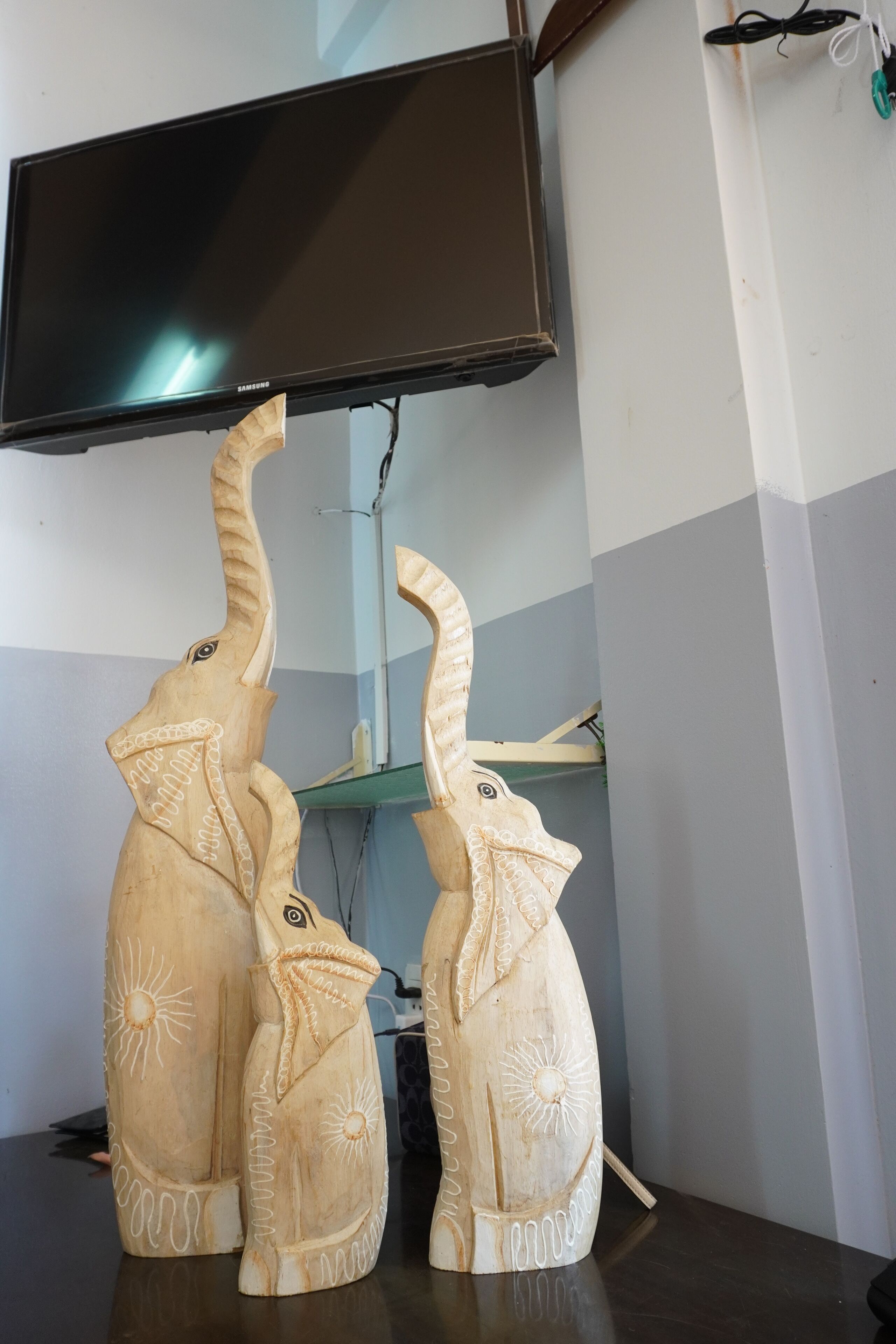 Deux sculptures d’éléphants en bois, finement sculptées, sont posées sur une surface sombre, et un téléviseur est fixé au-dessus.