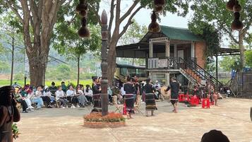 An outdoor gathering with people seated and standing, a building with a green roof, and a tree in the foreground.