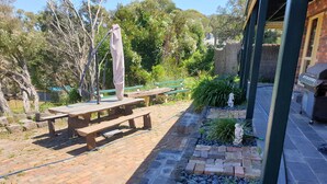Un espace patio avec une table en bois et des bancs, entouré d’un jardin agrémenté de plantes et d’un sentier en pierre.