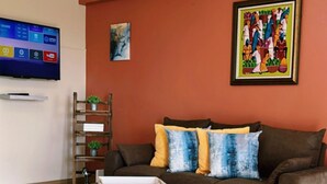 A living room with a brown sofa, a coffee table, a TV mounted on the wall, and a colorful painting above the sofa. - The Hideaway cozy , centrally located apartment 3 minutes from town center (Mandeville)
