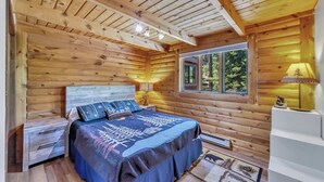 A log cabin bedroom with a bed, bedside lamps, a window, and a small table with a lamp.