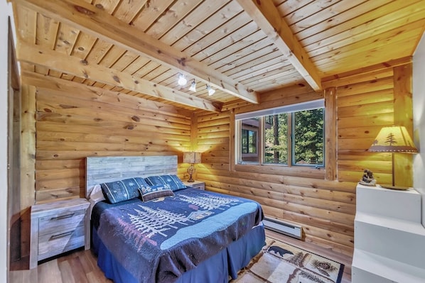 Un dormitorio tipo cabaña de troncos con una cama, lámparas de noche, una ventana y una mesita con lámpara.