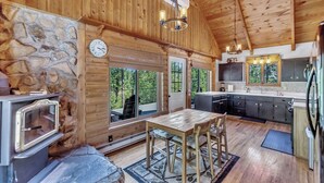 A cabin interior with a stone wall, wooden ceiling, a dining area, kitchen, and large windows.
