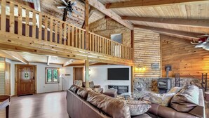 A spacious living room with a high ceiling, wooden beams, a stone fireplace, and a large TV.