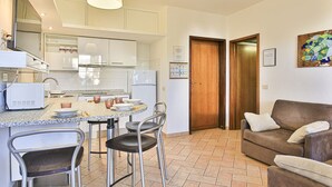 Una cocina moderna con armarios blancos, encimera de granito y un comedor con sillas de metal. Hay un sofá de cuero marrón y una puerta de madera en la sala de estar.