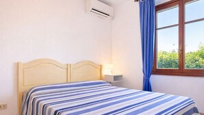 Un dormitorio con colcha a rayas, una mesita de noche con lámpara, un equipo de aire acondicionado y una ventana con cortinas.