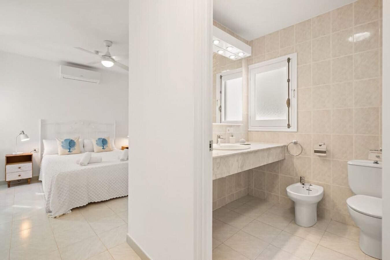 A modern hotel room with a neatly made bed, a bedside table with a lamp, a ceiling fan, and a bathroom with a sink, toilet, and bidet.