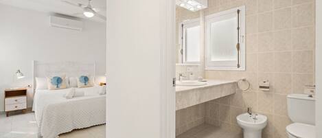 A modern hotel room with a neatly made bed, a bedside table with a lamp, a ceiling fan, and a bathroom with a sink, toilet, and bidet.