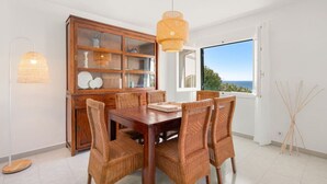 A dining area with a wooden table and wicker chairs, a glass cabinet with decorative items, and a view of the sea through a large window. - Villa Calma a gift for your eyes (San Jaime Mediterráneo)