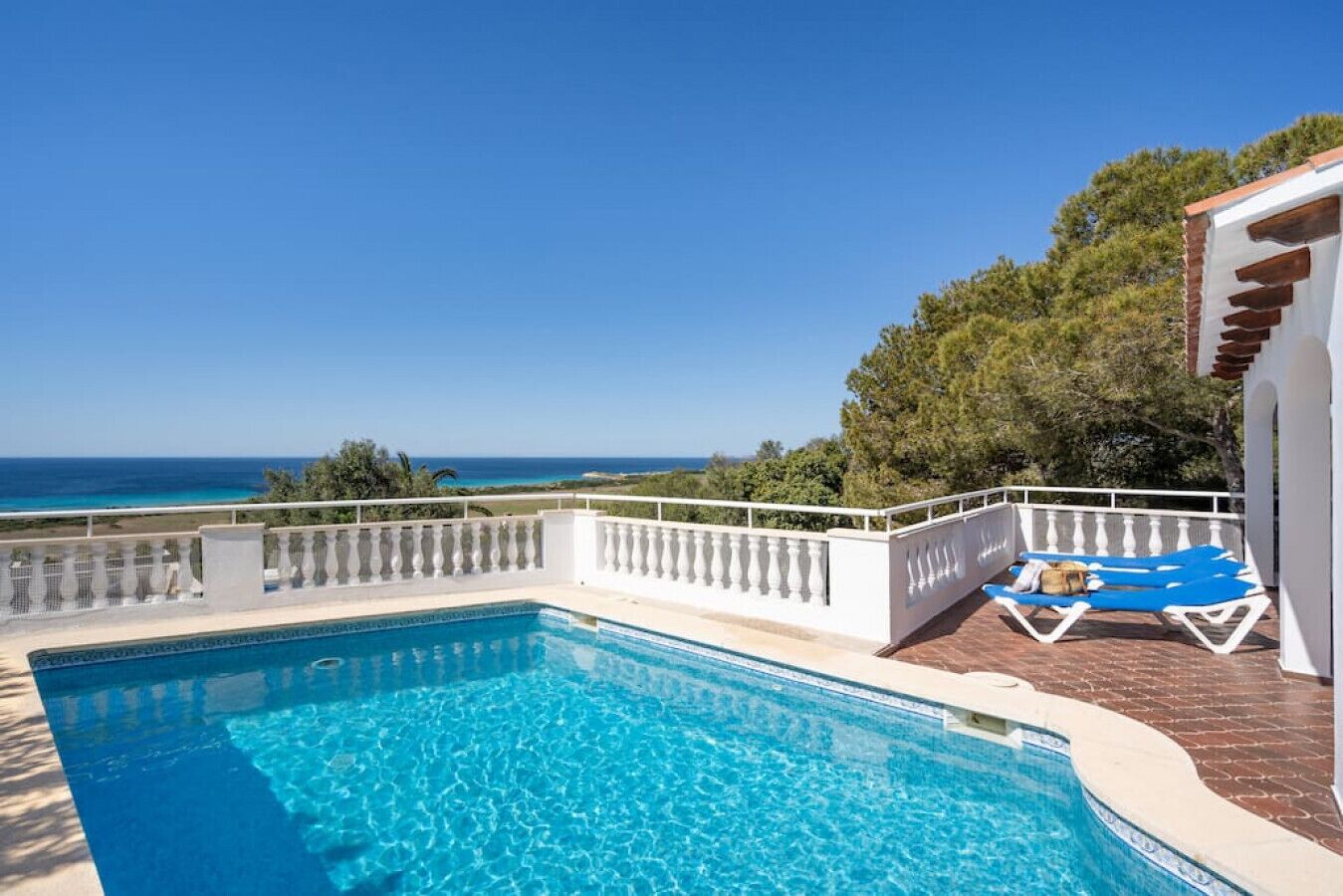 A swimming pool with a view of the sea, a white fence, and a blue lounge chair.