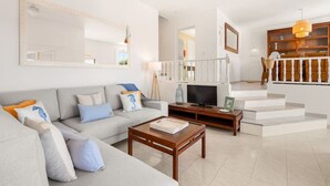 A modern living room with a grey sofa, wooden coffee table, and a flat-screen TV. A staircase with a wooden railing leads to an upper floor. - Villa Calma a gift for your eyes (San Jaime Mediterráneo)