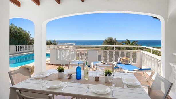 A dining area with a view of the sea, a pool, and a white building with arches. - Villa Calma a gift for your eyes (San Jaime Mediterráneo)
