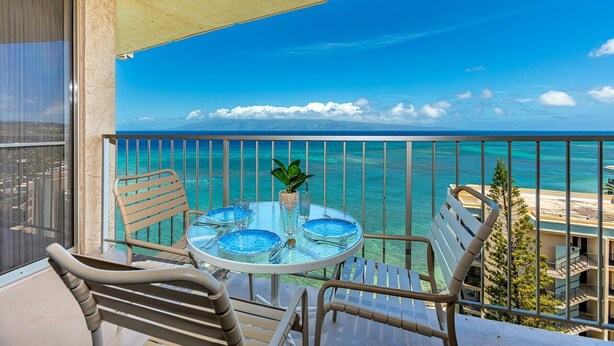 A balcony with a table set for two, overlooking a clear blue sea and distant land.