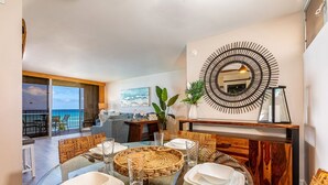 A dining area with a glass table, white plates, and wicker baskets. There is a large mirror on the wall and a view of the ocean through large windows.