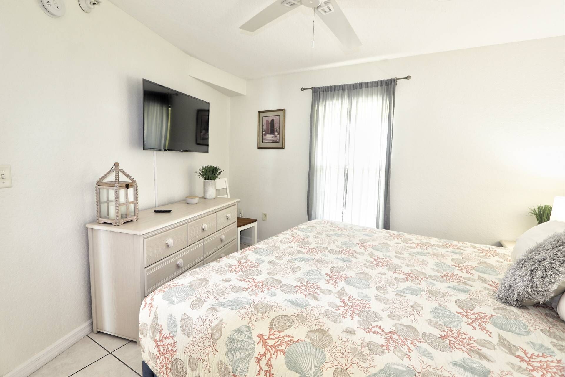 A bedroom with a bed, a dresser, a television mounted on the wall, and a window with curtains.
