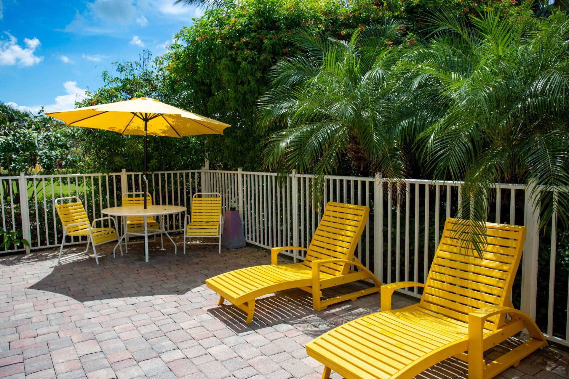 An outdoor patio area with yellow chairs and a table, surrounded by a white fence and lush greenery.