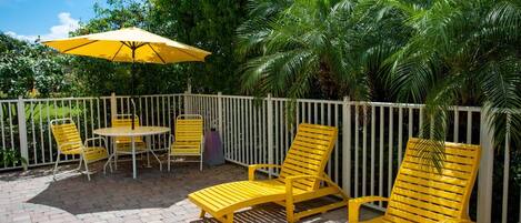 An outdoor patio area with yellow chairs and a table, surrounded by a white fence and lush greenery.