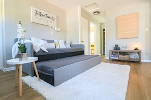 Ein modernes Schlafzimmer mit einer grauen Schlafcouch, einem weißen Teppich, einem kleinen Beistelltisch mit einer Pflanze und einer Wand mit einem Familien-Schild.