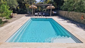 Une piscine rectangulaire bordée de pierres, entourée d’un mur de pierres et agrémentée d’une pergola surmontée de parasols en chaume.