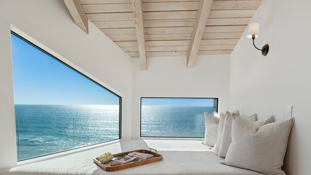 Ein Zimmer mit großem Fenster mit Meerblick, Holzboden und einem Bett mit Kissen sowie einem Tablett mit Gegenständen.