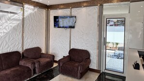 A modern waiting area with two plush armchairs, a ceiling fan, a mounted TV, and a view of the outside through a glass door. - Hotel Orchid Inn - Andheri (Mumbai)