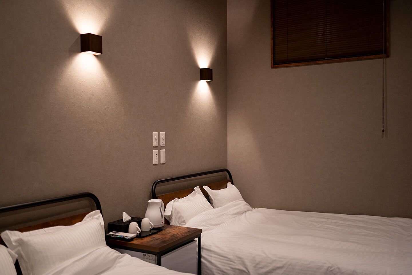 Hotellihuone, jossa on kaksi sänkyä, yöpöydät, seinään kiinnitetty valaisin ja ikkuna, jossa on sälekaihtimet.