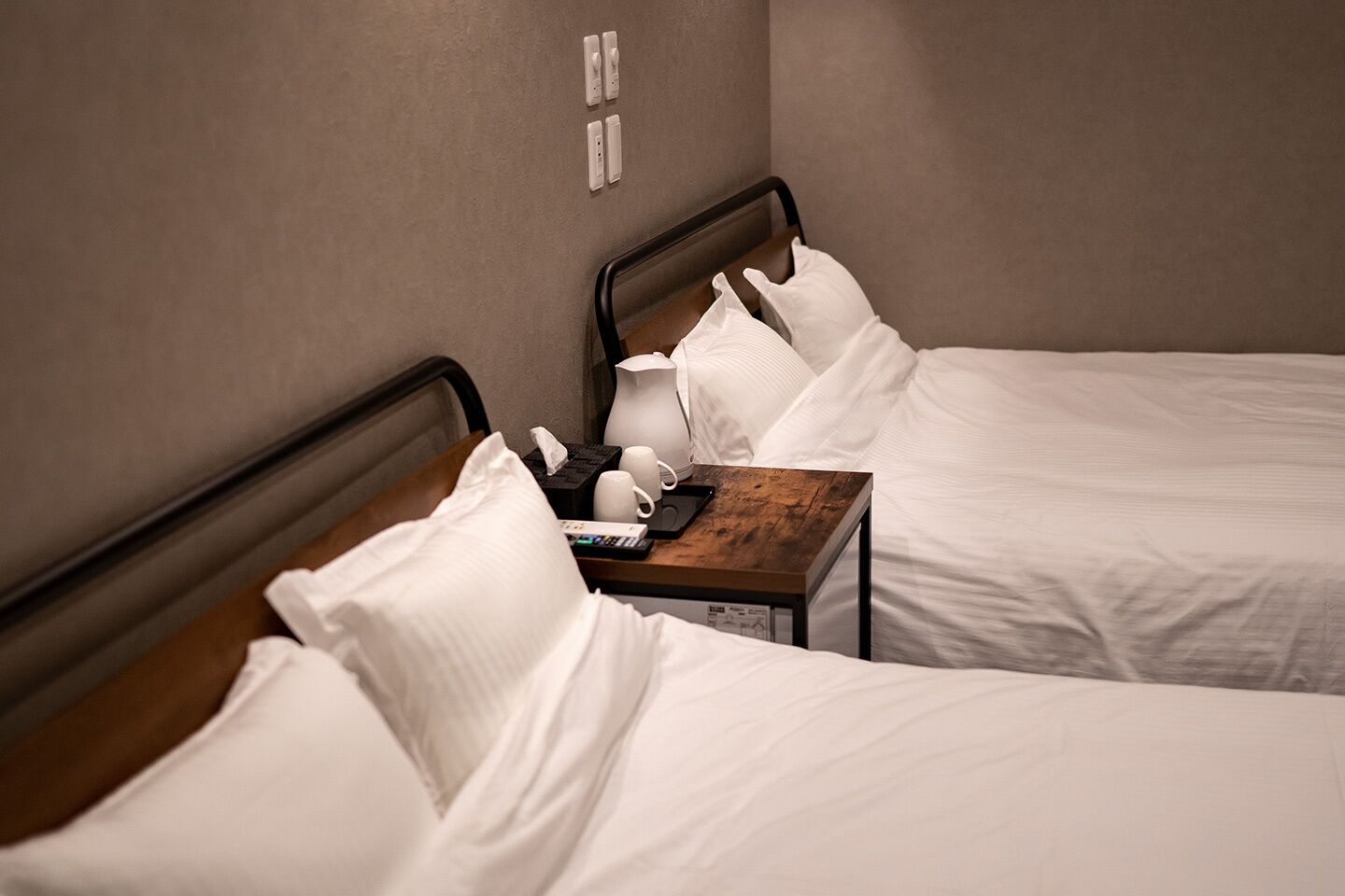 A hotel room with two beds, a wooden nightstand, and a wall-mounted power outlet.