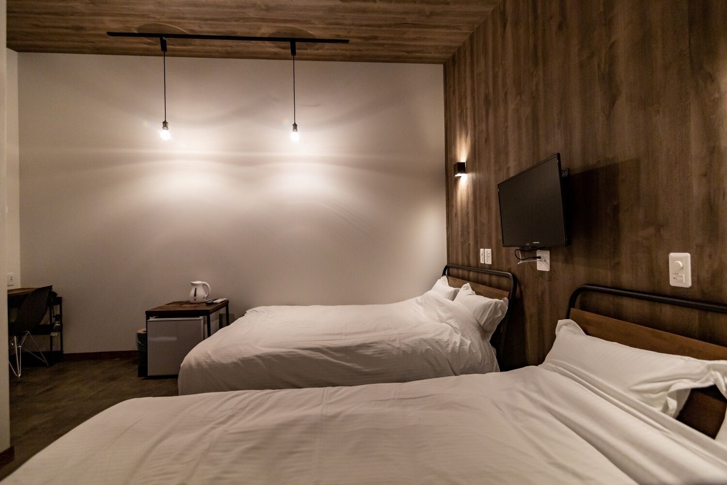 A hotel room with two beds, a TV mounted on the wall, a small table with a kettle, and a chair.
